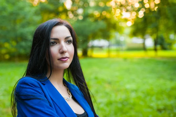 Hermosa morena en un traje azul en el fondo de la naturaleza. Descansa en el parque después del trabajo. Ojos marrones violentos . — Foto de Stock