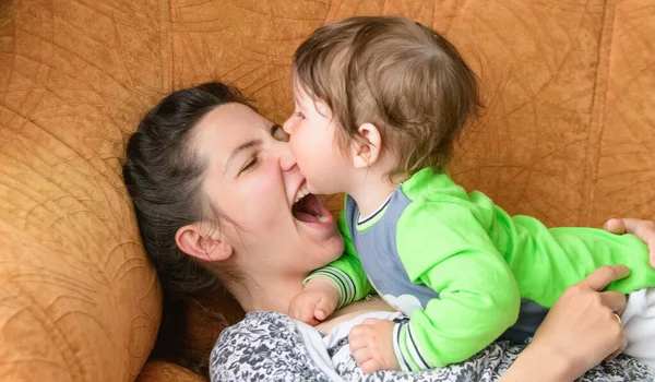 Ребенок кусает мамочку за нос. Счастливая мать с ребенком. Мальчик кусает матерей за нос на диване. Веселишься со своей любимой матерью — стоковое фото