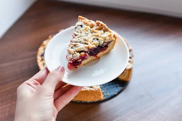 Tarta con bayas: frambuesas, fresas, grosellas, en un plato blanco, la mano de una mujer sosteniendo un pedazo de pastel en una espátula. Sobre un fondo de madera, en la servilleta de lino de fondo — Foto de Stock