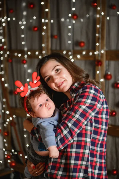 Mom and baby hug at the Christmas tree. Happy baby and his mom are looking at the frame. Mom with son hugging for Christmas. and have fun. newborn baby for the new year.