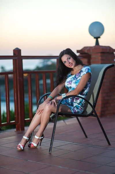Morena sentada en una silla al aire libre. Hermosa joven morena, vestida con un vestido corto y brillante, hermosa modelo femenina posando en la noche . — Foto de Stock