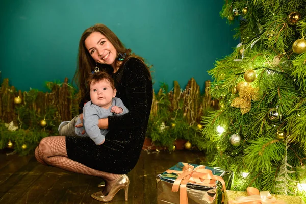 Mom and baby hug at the Christmas tree. Happy baby and his mom are looking at the frame. Mom with son hugging for Christmas. and have fun. newborn baby for the new year.