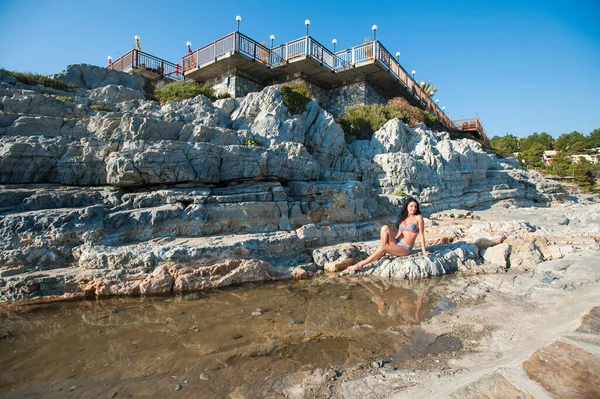 Una hermosa morena en bikini está sentada en una piedra. Sexy chica morena en bikini posando en una playa — Foto de Stock