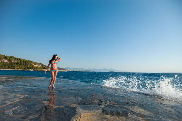 A girl in a swimsuit, splashing from the waves, Brunette girl rejoices splashing from the waves. Young happy woman jumping with raised arms, waving a blue bikini swimsuit in a blue swimsuit and a spla — Φωτογραφία Αρχείου