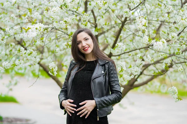 Schwangere Brünette im blühenden Park. die Brünette im dunklen Kleid posiert im blühenden Apfelgarten, das Kleid versteckt sich schwanger — Stockfoto