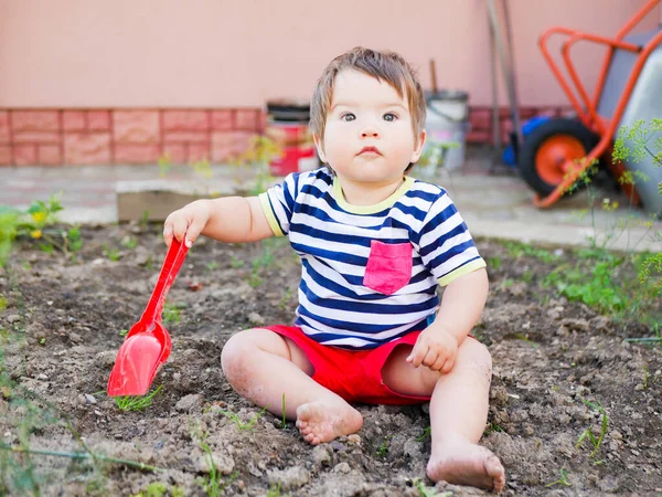 Ребенок играет с красным детским лопаткой. Маленький мальчик копается в земле — стоковое фото