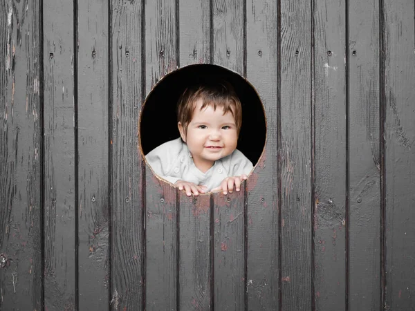 Kind kijkt door een rond gat. donkere houten achtergrond. glimlacht naar de camera. plaats voor inscripties. portret van gelukkig jongetje, het kind kijkt uit rond houten raam en glimlacht — Stockfoto