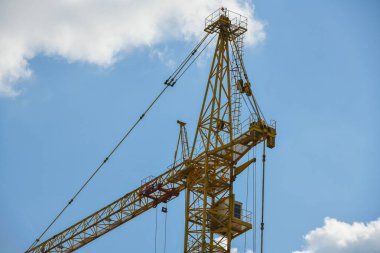 Yapı aşamasındaki beton ve tuğladan oluşan çok katlı yeni bir binanın üzerinde mavi gökyüzüne karşı uzun sarı oklu bir vinç.