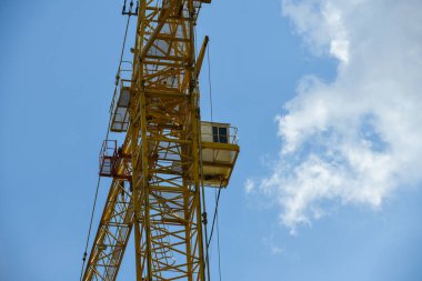 Yapı aşamasındaki beton ve tuğladan oluşan çok katlı yeni bir binanın üzerinde mavi gökyüzüne karşı uzun sarı oklu bir vinç.