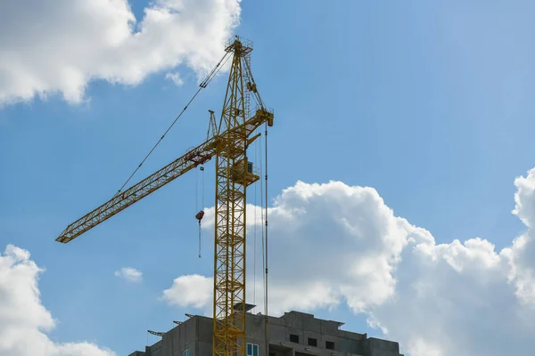 High-rise construction crane with a long arrow of yellow color against the blue sky over a new multi-storey building of concrete and brick under construction — ストック写真