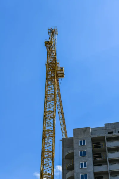 High-rise construction crane with a long arrow of yellow color against the blue sky over a new multi-storey building of concrete and brick under construction — ストック写真