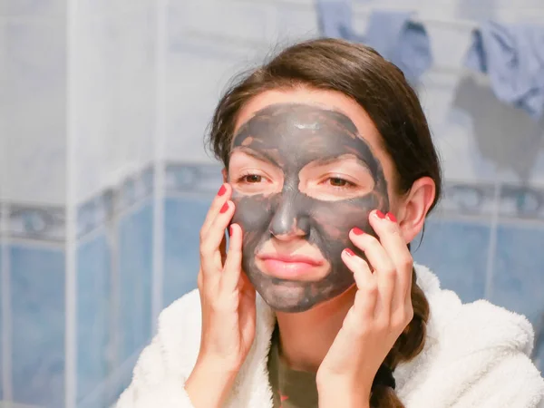 The girl in the face mask looks in the mirror displeased. Cosmetic black mask. Woman brings beauty to her face, gets rid of black dots, using a black mask. A female in the bathroom looks in the mirror — Stock Photo, Image
