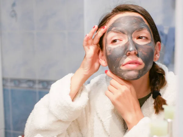 The girl is caring for the face. Cosmetic black mask. A woman brings beauty to her face, gets rid of black dots with a black mask. Woman in the bathroom looks in the mirror — Stock Photo, Image