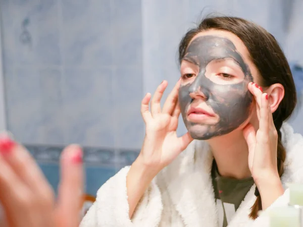 The girl is caring for the face. Cosmetic black mask. A woman brings beauty to her face, gets rid of black dots with a black mask. Woman in the bathroom looks in the mirror — Stock Photo, Image