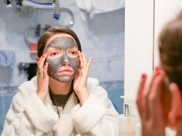 The girl is caring for the face. Cosmetic black mask. A woman brings beauty to her face, gets rid of black dots with a black mask. Woman in the bathroom looks in the mirror — Stock Photo, Image