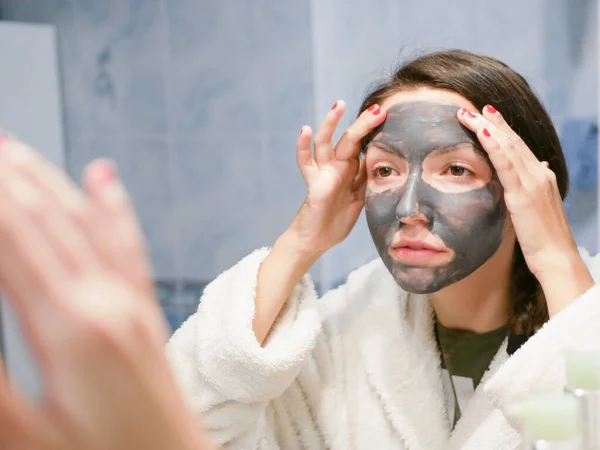 the girl in the face mask looks in the mirror displeased. Cosmetic black mask. Woman brings beauty to her face, gets rid of black dots, using a black mask. A female in the bathroom looks in the mirror