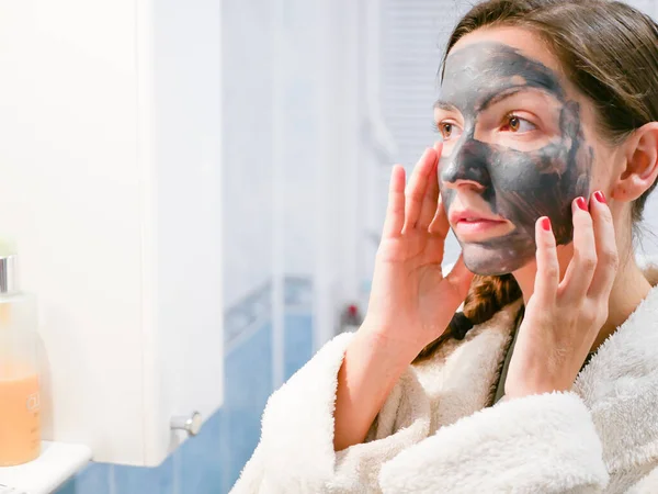 The girl is caring for the face. Cosmetic black mask. A woman brings beauty to her face, gets rid of black dots with a black mask. Woman in the bathroom looks in the mirror — Stock Photo, Image