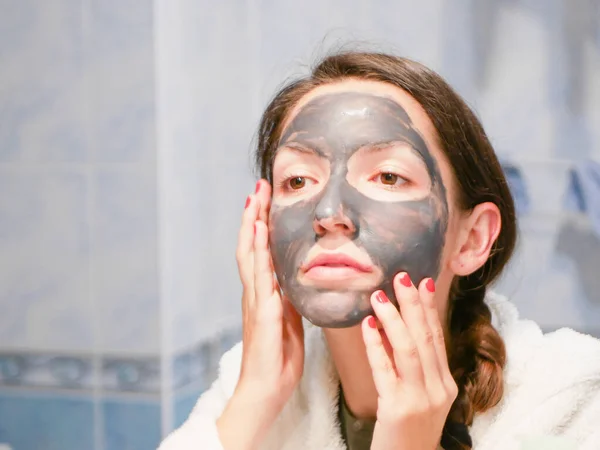 Máscara preta cosmética. Mulher traz beleza para o rosto, se livra de pontos pretos, usando uma máscara preta. Uma fêmea no banheiro olha no espelho — Fotografia de Stock