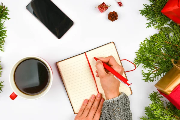 Wishes for next year. Woman hand on the background of fir branches. New Year\'s winter holiday. To-do list on a notepad next to Christmas decorations, pine cones and a coffee mug. Layout concept, New Y