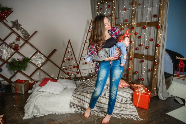 Mom and baby hug at the Christmas tree. Happy baby and his mom are looking at the frame. Mom with son hugging for Christmas. and have fun. newborn baby for the new year.