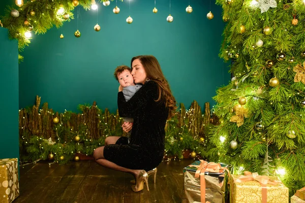 Mom and baby hug at the Christmas tree. Happy baby and his mom are looking at the frame. Mom with son hugging for Christmas. and have fun. newborn baby for the new year. — Stock Photo, Image