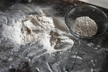 Masasında un olan Colander. Paslanmaz çelik ince ağlı süzgeç süzgeci un süzgeci, koyu renk arka planda un ile kaplanmış. Mutfak eşyaları..