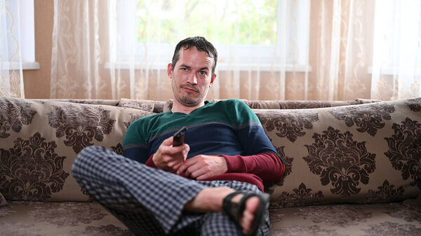 Недовольный мужчина сидит на диване с пультом от телевизора.