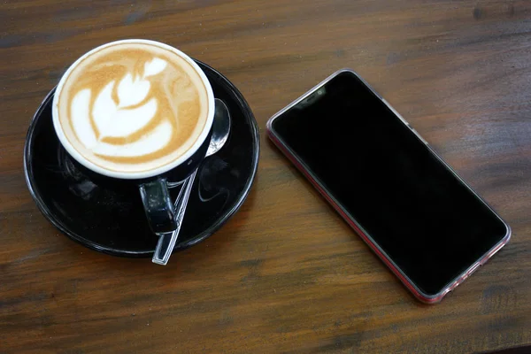 Filiżanka kawy z pięknym latte sztuki na drewnianym stole bar obok telefonu komórkowego — Zdjęcie stockowe