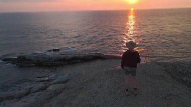 Kamera, uçurumun kenarında güneşin batışıyla Newfoundland, Cape St. George 'da duran kadının etrafında dönüyor..