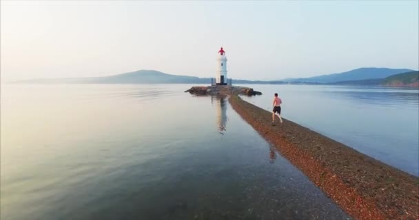 Vue aérienne d'un jeune homme en short courant rapidement le long de la flèche Tokarevsky vers le phare. La caméra suit le sportif. Belle mer du matin au lever du soleil. Vladivostok, Russie 