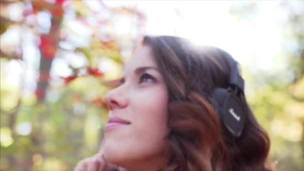 ヘッドフォンで音楽を聴く 秋の公園の素晴らしい日光を楽しんで カメラに笑顔のきれいな女性の近い肖像画 — ストック動画