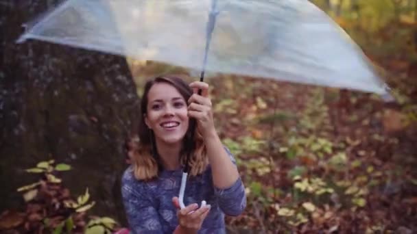 毛布の上にツリーの下に座って透明傘で遊んで笑顔の若い女性の肖像画 秋の森 — ストック動画