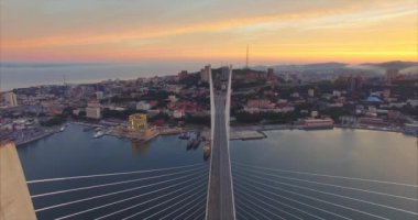 Büyük Altın Köprü sütunları arasında uçmak, Golden Horn körfezi boyunca köprünün havadan manzarası, yol, güzel güneş doğarken Vladivostok şehri. Yaz. Rusya