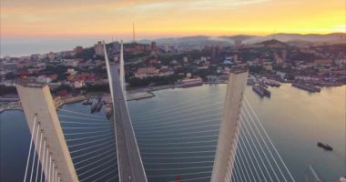 Golden Bridge sütunları arasında uçmak, Golden Horn körfezi üzerindeki köprünün hava manzarası, sürüş araçlarıyla yol, güzel güneş doğarken Vladivostok şehri manzarası. Yaz. Rusya
