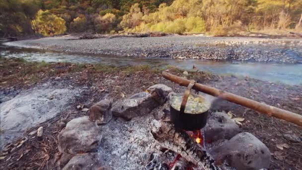キャンプファイヤーの上の鍋の景色を背景に流れる川の近くの新鮮な魚からおいしい魚のスープ — ストック動画