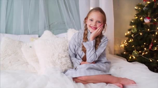 大きなベッドの上に座ってカメラに笑みを浮かべて2つの編組とかわいい女の子のスライド肖像画 クリスマスツリーが背景にあります — ストック動画