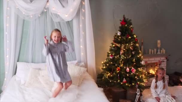 两个辫子的可爱的快乐的女孩跳到床上 笑着玩着 圣诞树和壁炉后面是另一个女孩 — 图库视频影像