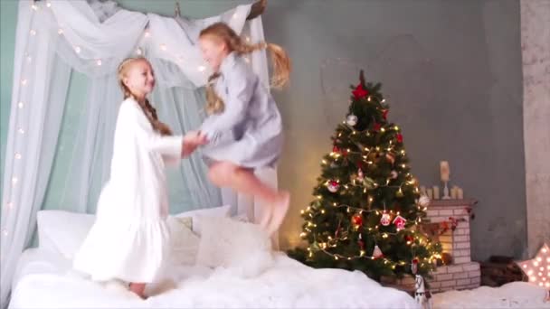 两个光着脚 手牵手 手牵着辫子 高高地跳在床上的女孩的慢镜头 萤火虫和圣诞树在后面 — 图库视频影像