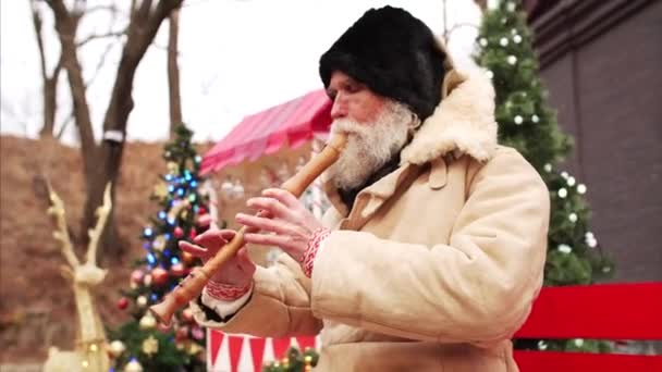 暖かいコートと黒の帽子の白い髭の老人の肖像は クリスマスの装飾やクリスマスツリーの間でベンチで木製のパイプを演奏 — ストック動画