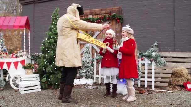 在圣诞节展销会上 身穿大衣戴帽子的白胡子老人正在给两个穿着红色外套的女孩自制饼干 — 图库视频影像