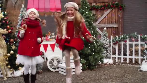 赤いコートの二つのかわいい笑顔の女の子はクリスマスフェアの装飾の中で高くジャンプしている — ストック動画