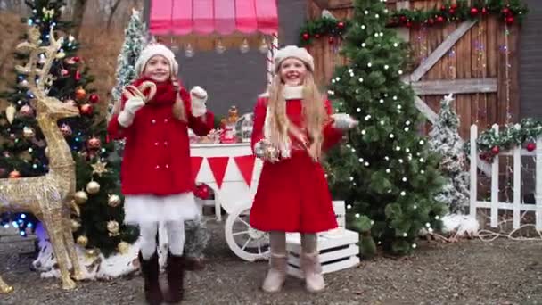赤いコートの二人のかわいい笑顔の女の子はクリスマスフェアの装飾の間でジャンプしている — ストック動画
