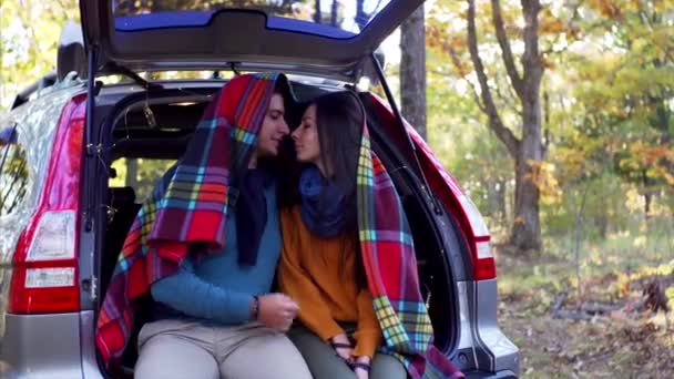 年轻的男人和女人在温暖的格子花下 坐在汽车行李箱里开心地笑着 阳光明媚的秋木 — 图库视频影像