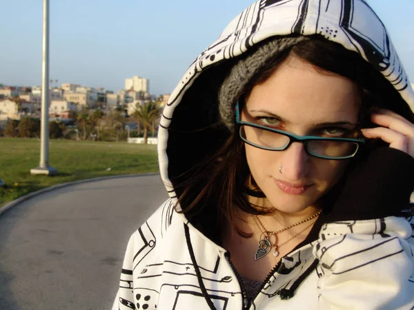 Young adult female starring at the camera through blue glasses, wearing a casual hat and a hoodie, urban style.