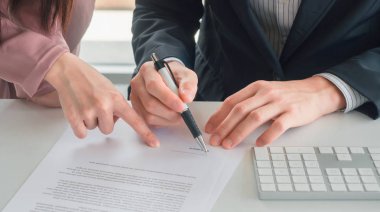 Pembe takım elbiseli iş kadını, masanın üzerinde imza için lacivert takım elbiseli işadamına belge gönderiyor. Belgedeki imza pozisyonu için parmak noktasını kullanıyor ve o da kalem tutuyor. İş kavramı.