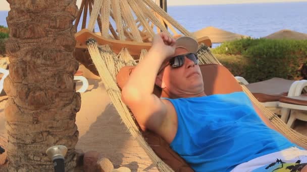 在埃及红海岸边 一个阳光明媚的夏日 一个头戴黑色帽子 戴着太阳镜的年轻人躺在吊床上 躺在棕榈树树阴下 — 图库视频影像