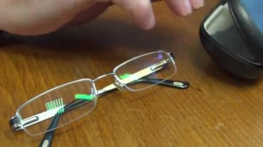 Erkek eli siyah bir kutudan gözlük çıkarır ve cam lensi toz ve pislikten bir bezle siler. Yakın plan.