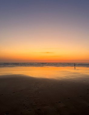 Children play on the sandy shore of Biscarosse as the sun sets, painting the sky in warm hues. The calm waves gently lap at their feet, creating a peaceful atmosphere.