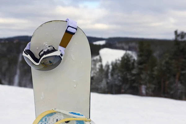 a snowboard stuck in the snow and ski goggles hanging on it. Concept to illustrate ski admission fee.