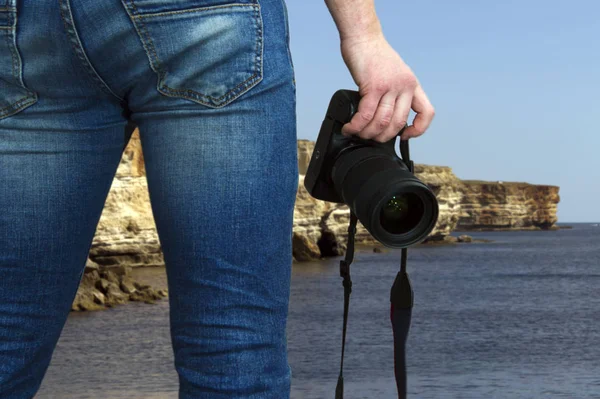 View of the back of a man standing in front of the sea and holding a digital camera in his hands, copy space
